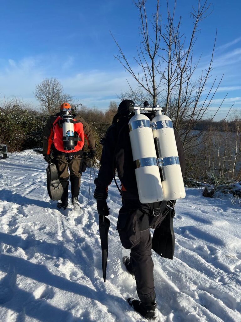 Bild zweier Gerätetaucher, die durch eine Schneelandschaft zum Einstig des Sees wandern.