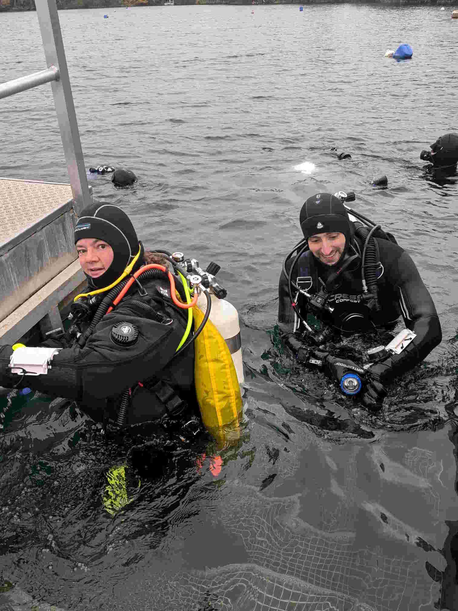 Bild zweier Gerätetaucher, die lächelnd an einer Leiter herunter in einen See absteigen.
